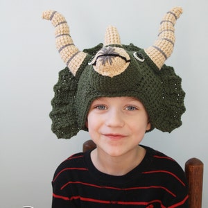 Puede incluir: Un ni&ntilde;o con un gorro de triceratops de crochet verde, marr&oacute;n y beige. El gorro tiene una cabeza grande y redonda con dos cuernos y una gola. El ni&ntilde;o sonr&iacute;e y mira a la c&aacute;mara.