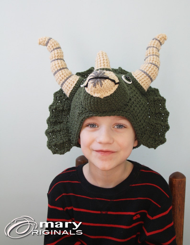 Puede incluir: Un ni&ntilde;o con un gorro de triceratops de crochet verde, marr&oacute;n y beige. El gorro tiene una cabeza grande y redonda con dos cuernos y una gola. El ni&ntilde;o sonr&iacute;e y mira a la c&aacute;mara.