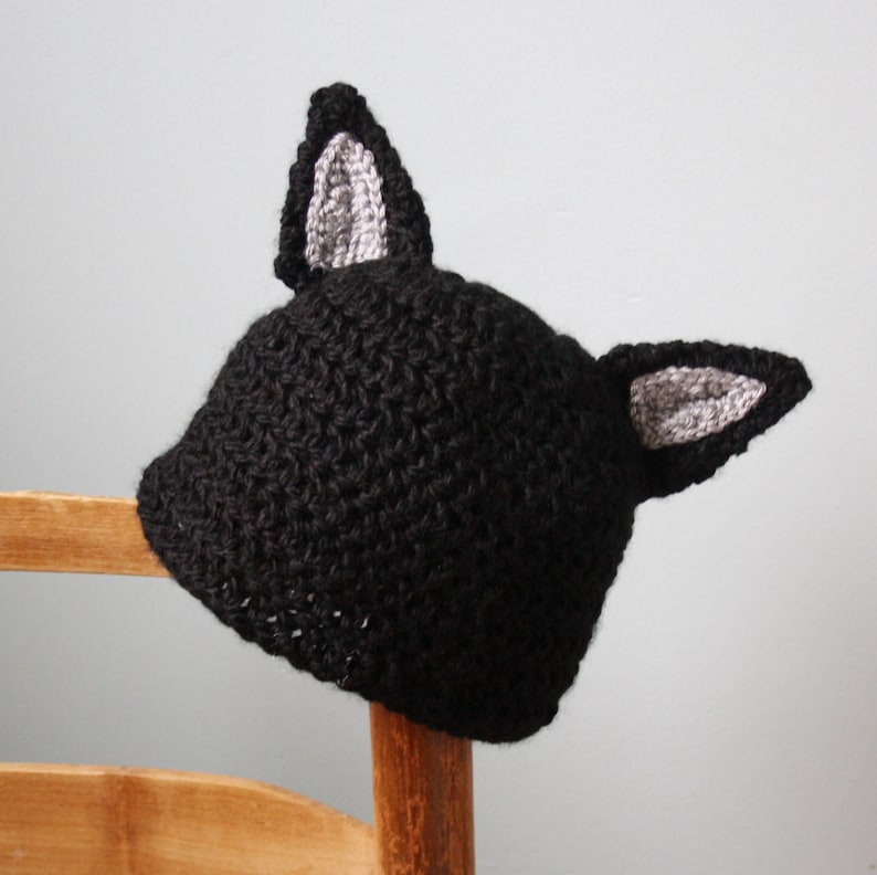 May include: Black crocheted hat with gray cat ears. The hat is sitting on a wooden chair.