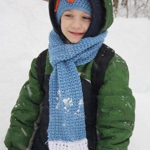 May include: A young person wearing a green jacket, blue scarf, and a knitted hat with an orange flower design. The scarf has a white and gray stripe at the bottom. The person is standing in a snowy environment.