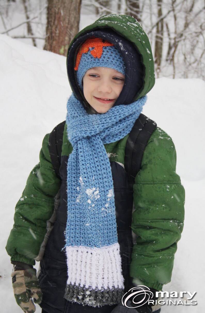 May include: A young person wearing a green jacket, blue scarf, and a knitted hat with an orange flower design. The scarf has a white and gray stripe at the bottom. The person is standing in a snowy environment.