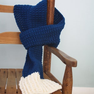 May include: A blue and white crocheted scarf draped over a wooden chair. The scarf is made with a textured stitch and has a long, flowing design.