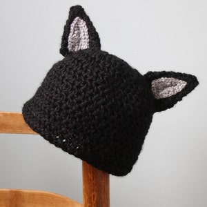 May include: Black crocheted hat with gray cat ears. The hat is sitting on a wooden chair.