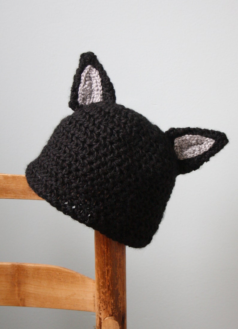 May include: Black crocheted hat with gray cat ears. The hat is sitting on a wooden chair.