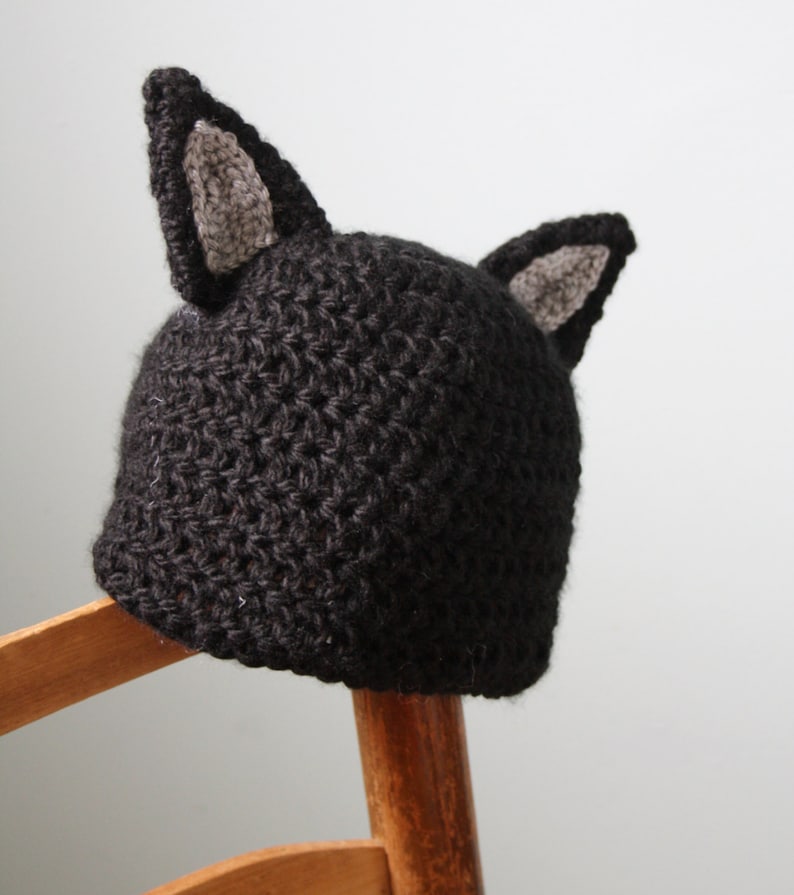 May include: A black crocheted hat with gray cat ears. The hat is sitting on a wooden chair.