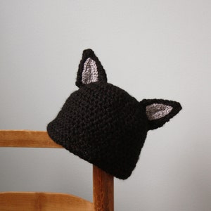 May include: Black crocheted hat with gray crocheted ears. The hat is sitting on a wooden chair.