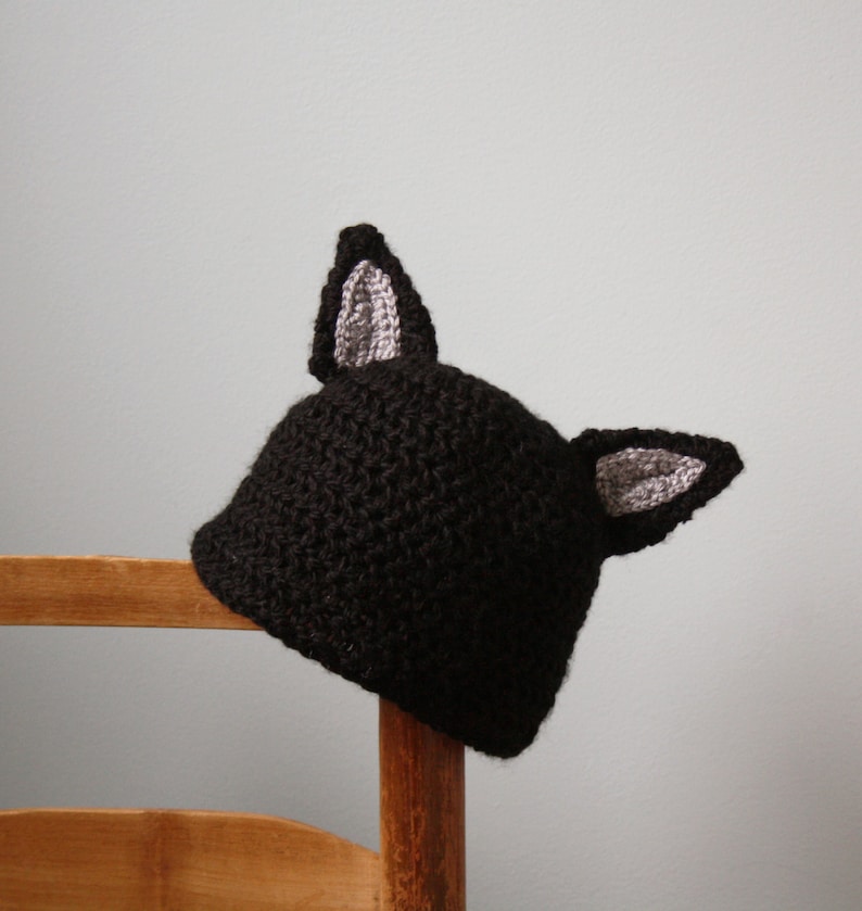 May include: Black crocheted hat with gray crocheted ears. The hat is sitting on a wooden chair.