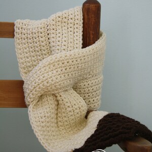 May include: A crocheted scarf in cream and brown with a knotted design. The scarf is draped over a wooden chair.