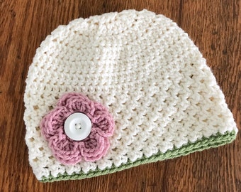 May include: A white crocheted beanie with a green crocheted brim and a pink crocheted flower with a white button in the center.