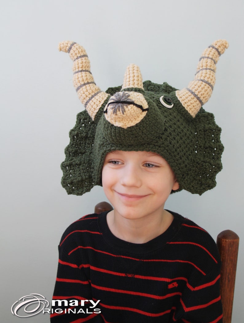 Puede incluir: Un ni&ntilde;o con un gorro de triceratops de color verde tejido a crochet con cuernos beige y ojos de bot&oacute;n negros. El gorro tiene un volante de crochet alrededor de la parte inferior. El ni&ntilde;o lleva una camisa a rayas negras y rojas.
