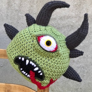 May include: A handmade, green crocheted monster hat with a large, red-rimmed eye, black horns, and a wide, toothy mouth. The hat features a red tongue and is designed to resemble a cartoonish, scary creature. The hat is displayed on a wooden post.