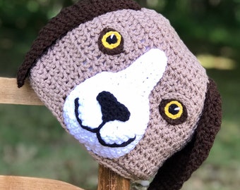 May include: A brown and white crocheted dog hat with black ears and yellow eyes. The hat is sitting on a wooden chair.