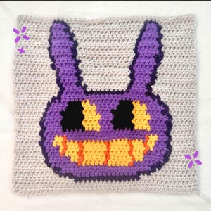 May include: A crocheted square featuring a purple cartoon character with large ears, black eyes, and an orange mouth. The character is set against a light gray crocheted background. The square is a handmade item.