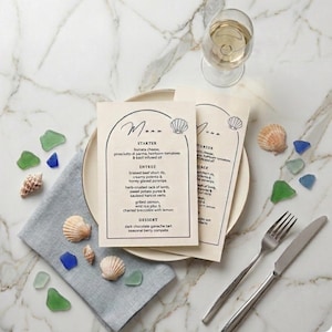 May include: A restaurant table setting featuring two menu cards with a seashell design, a wine glass, and cutlery. Seashells and sea glass in green and blue are scattered around the plates.