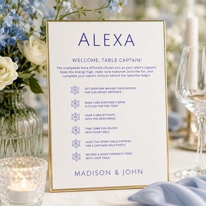 Könnte beinhalten: Ein rechteckiges weißes Schild in einem dünnen goldenen Rahmen mit blauem Text, der ALEXA, WELCOME, TABLE CAPTAIN! lautet, gefolgt von einer Liste von Hochzeitsaufgaben mit Steuerrad-Symbolen und den Namen MADISON & JOHN am unteren Rand.