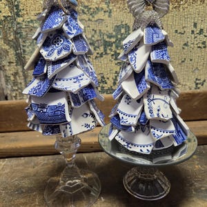 May include: Two decorative Christmas trees crafted from blue and white china shards. One tree is mounted on a clear glass stem, the other on a mirrored base. The trees are topped with a dragonfly and a silver bow.