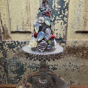 May include: A decorative Christmas tree made of silver-coloured materials and adorned with various jewels, pearls, and a crown on top. The tree sits on a clear glass stand with a decorative base, adding a touch of elegance to the festive design.