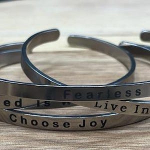 May include: Four silver-coloured metal cuff bracelets with engraved inspirational quotes. The quotes include "All I Need is", "Fearless", "Choose Joy", and "Live In The Moment". The bracelets are displayed on a wooden surface.
