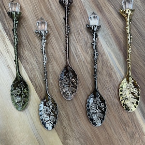 Puede incluir: Cinco cucharas adornadas con tapas de cristal. Las cucharas tienen un diseño de hoja y están en diferentes acabados metálicos: oro, plata, bronce y verde.