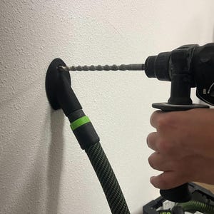 Puede incluir: Un taladro negro y verde con una manguera de extracción de polvo negra y verde conectada al taladro. El taladro se está utilizando para perforar un agujero en una pared blanca.
