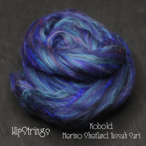 Puede incluir: Primer plano de una bola de fibra de merino, shetland y tussah sari azul, verde y negro. La fibra está dispuesta en un patrón suelto y en espiral. El texto "HipStrings Kobold Merino Shetland Tussah Sari" es visible en la parte inferior de la imagen.