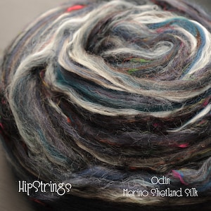 Puede incluir: Primer plano de un ovillo de lana en tonos grises, blancos y azules con motas de colores. La lana está etiquetada como "Odin Merino Shetland Silk" y "HipStrings88".