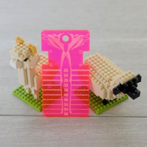 May include: A bright pink acrylic knitting needle gauge tool with a floral cutout at the top and numerical markings from 20 to 80, positioned between two small beige brick-built animal figures standing on green square base plates.