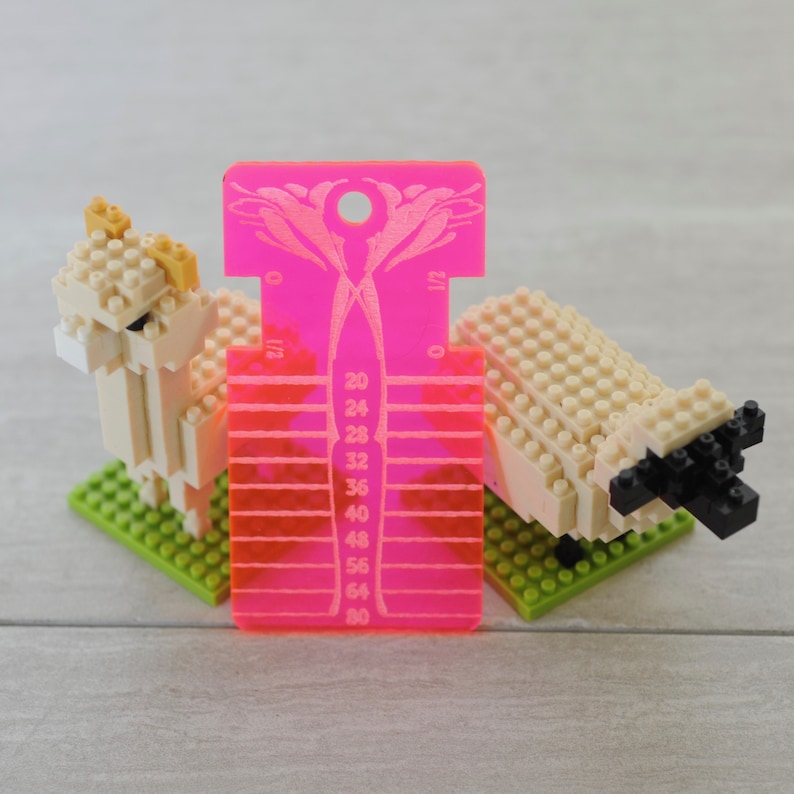 May include: A bright pink acrylic knitting needle gauge tool with a floral cutout at the top and numerical markings from 20 to 80, positioned between two small beige brick-built animal figures standing on green square base plates.