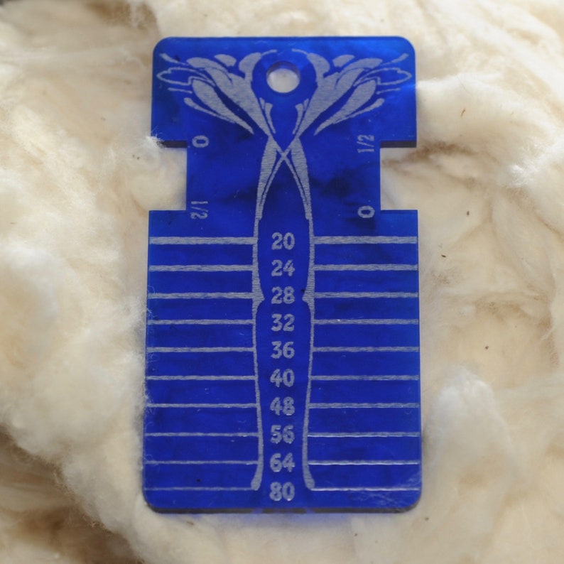 May include: A rectangular blue acrylic knitting needle gauge tool featuring a central column of numbers ranging from 20 to 80, with horizontal measurement lines extending to the sides and a decorative floral-style cutout at the top with a hole.