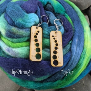 May include: A pair of wooden needle gauge earrings with silver hooks. The gauges are marked with sizes from 00 to 10.5. The earrings are labeled "HipStrings" and "Maple".