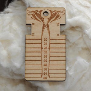 May include: A light-colored wooden yarn gauge tool featuring a central vertical column with numerical markings from 20 to 80, horizontal slots on the sides, and a circular hole at the top, decorated with an engraved symmetrical floral pattern.