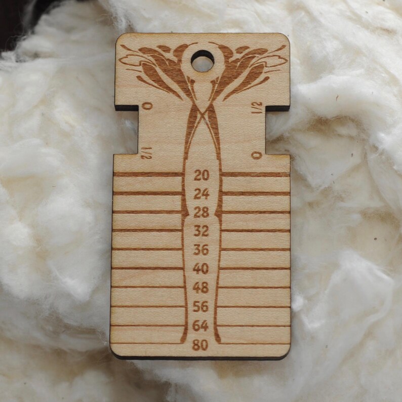 May include: A light-colored wooden yarn gauge tool featuring a central vertical column with numerical markings from 20 to 80, horizontal slots on the sides, and a circular hole at the top, decorated with an engraved symmetrical floral pattern.