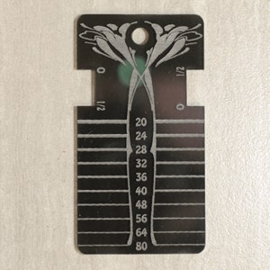 May include: A black rectangular gauge tool featuring a central vertical column with numerical markings 20, 24, 28, 32, 36, 40, 48, 56, 64, and 80. The tool includes horizontal lines, a circular hole at the top, and engraved floral-like patterns.