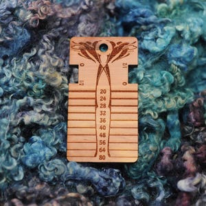 May include: A rectangular wooden yarn gauge tool featuring a laser-etched floral design at the top, a circular hole, and horizontal slots labeled with numbers 20, 24, 28, 32, 36, 40, 48, 56, 64, and 80, with 0 and 1/2 markings on the sides.
