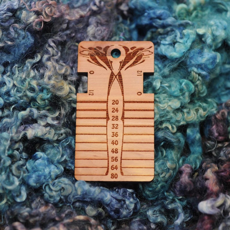 May include: A rectangular wooden yarn gauge tool featuring a laser-etched floral design at the top, a circular hole, and horizontal slots labeled with numbers 20, 24, 28, 32, 36, 40, 48, 56, 64, and 80, with 0 and 1/2 markings on the sides.
