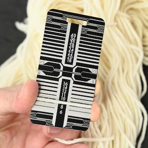 May include: A black metal gauge with a white yarn wrapped around it. The gauge has markings for measuring yarn thickness in inches. The markings range from 6 to 40.