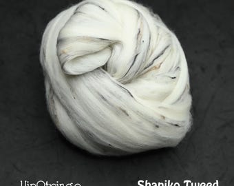 Puede incluir: Una bola de lana Shaniko Tweed. La lana es principalmente blanca con sutiles motas negras y marrones. El logotipo de HipStrings y el nombre del producto son visibles.