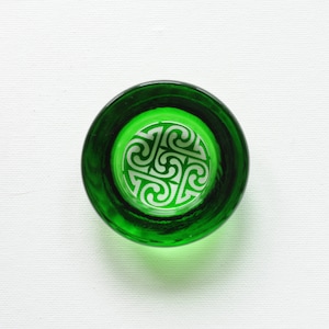 May include: A green glass dish with a white Celtic knot design. The dish is round and has a slightly raised rim.