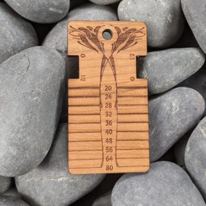 May include: A rectangular wooden knitting needle gauge tool featuring a laser-engraved floral design at the top, a circular hole for hanging, and horizontal slots labeled with numerical sizes ranging from 20 to 80, with additional 0 and 1/2 markings.