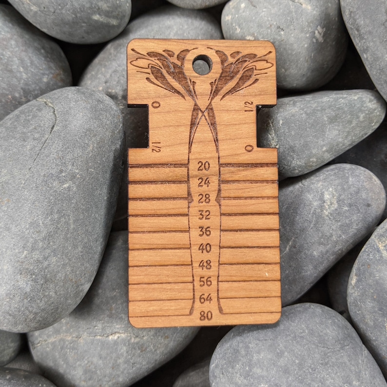 May include: A rectangular wooden knitting needle gauge tool featuring a laser-engraved floral design at the top, a circular hole for hanging, and horizontal slots labeled with numerical sizes ranging from 20 to 80, with additional 0 and 1/2 markings.