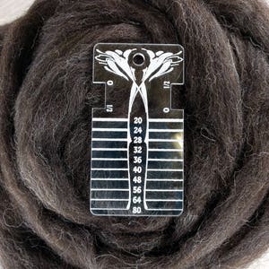 Puede incluir: Un primer plano de una lana virgen marrón con una galga de plástico transparente que tiene marcas de 20 a 80. La galga se utiliza para medir el diámetro de la fibra.