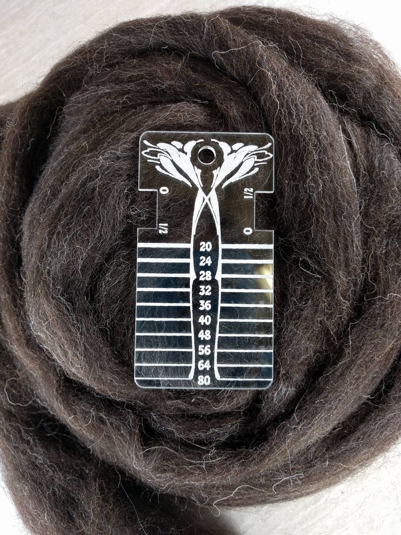 May include: A clear acrylic yarn gauge tool with white printed markings and numbers ranging from 20 to 80, featuring a decorative floral design at the top, resting on a coiled bundle of dark brown wool roving fiber.