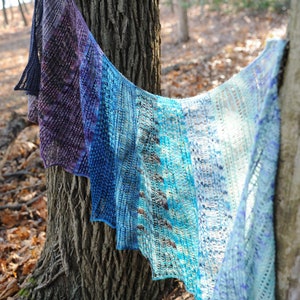 May include: A hand-knitted shawl in shades of purple, blue, and white, draped over a tree trunk in a woodland setting.