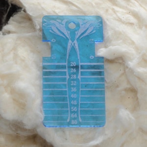 May include: A translucent blue acrylic yarn gauge tool featuring a central vertical column with numerical markings from 20 to 80, horizontal slots for measuring, and a circular hole at the top for hanging, with 0 and 1/2 inch markings.