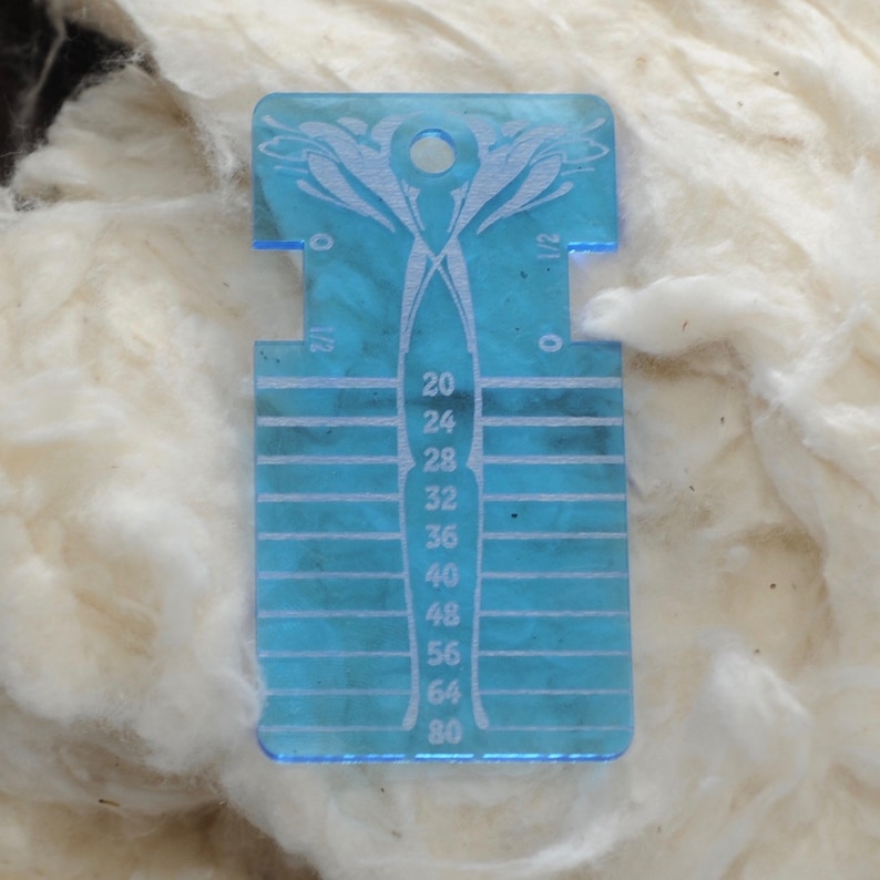 May include: A translucent blue acrylic yarn gauge tool featuring a central vertical column with numerical markings from 20 to 80, horizontal slots for measuring, and a circular hole at the top for hanging, with 0 and 1/2 inch markings.