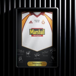 May include: A framed white soccer jersey with yellow and red accents. The jersey has the Adidas logo, the text "UEFA Cup Final 17 May 2000", and the text "Marshall boya". The jersey is signed by several players, including Taffarel, Hasan Sag, Gheorghe Hagi, and Jardel. The frame also includes the text "Galatasaray 1999-2000".