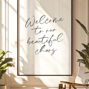Pode incluir: Uma impressão emoldurada com as palavras "Welcome to our beautiful chang" em elegante escrita cursiva. A moldura preta envolve um fundo branco. A imagem é exibida em uma sala bem iluminada com uma cadeira e plantas.
