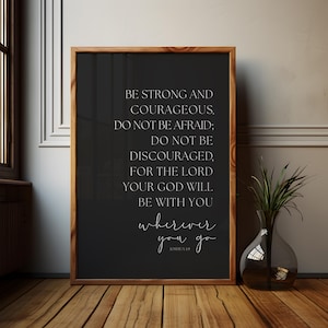 Könnte beinhalten: Ein gerahmter Druck mit Holzrahmen zeigt den Text "BE STRONG AND COURAGEOUS. DO NOT BE AFRAID; DO NOT BE DISCOURAGED, FOR THE LORD YOUR GOD WILL BE WITH YOU wherever you go." Eine dekorative Vase mit Grün steht daneben.