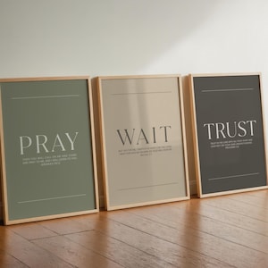 May include: Three framed art prints with inspirational words: "PRAY", "WAIT", and "TRUST". Each print has a neutral color background and a wooden frame. The text is in a simple, elegant font. The prints are leaning against a wall.