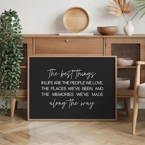 Op de afbeelding: Een zwart, ingelijst bord met witte tekst: "The best things in life are the people we love, the places we've been, and the memories we've made along the way". Het bord staat op een houten kast.